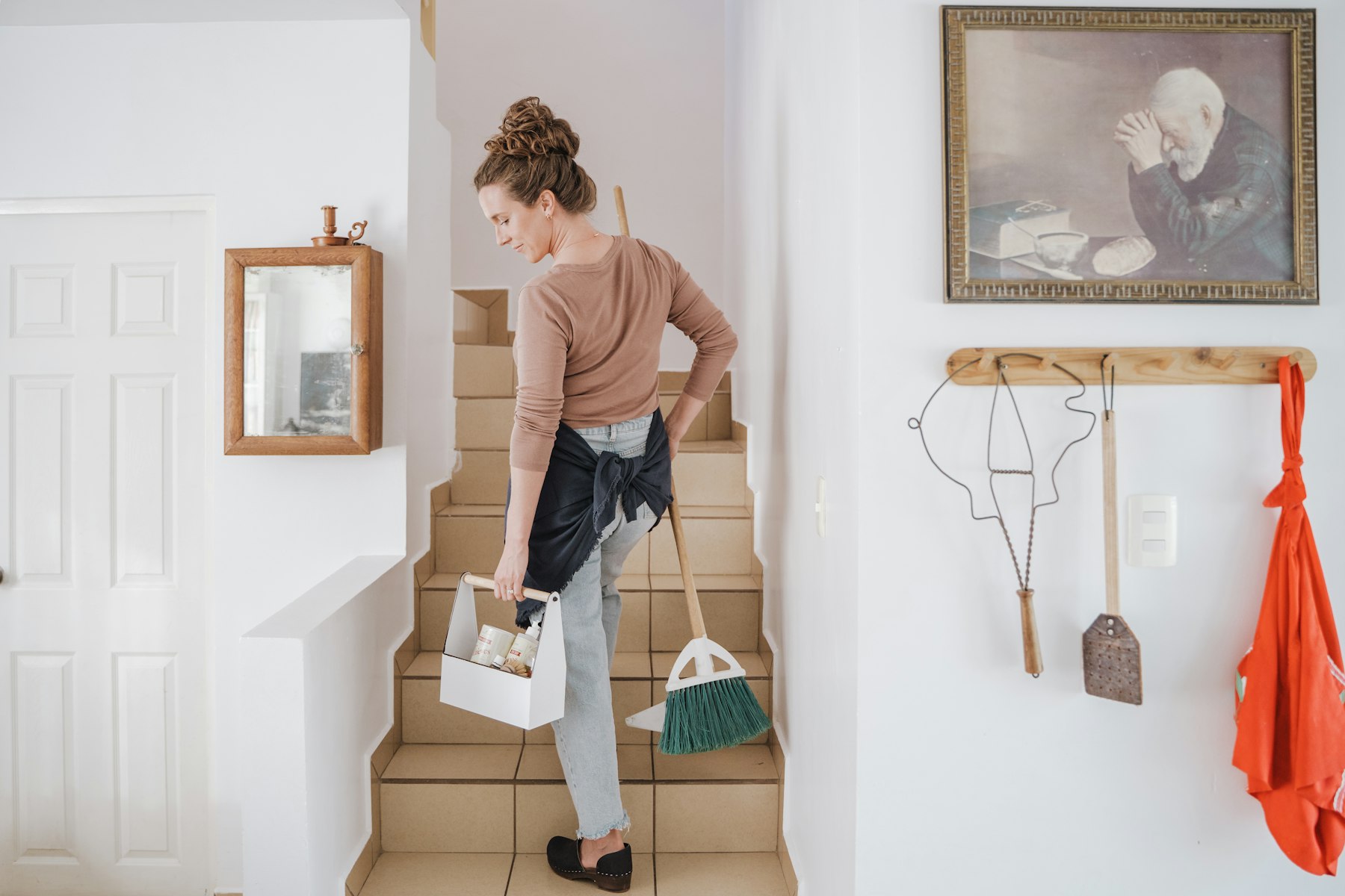 A cleaner carrying a caddy and broom up the stairs of a bright home